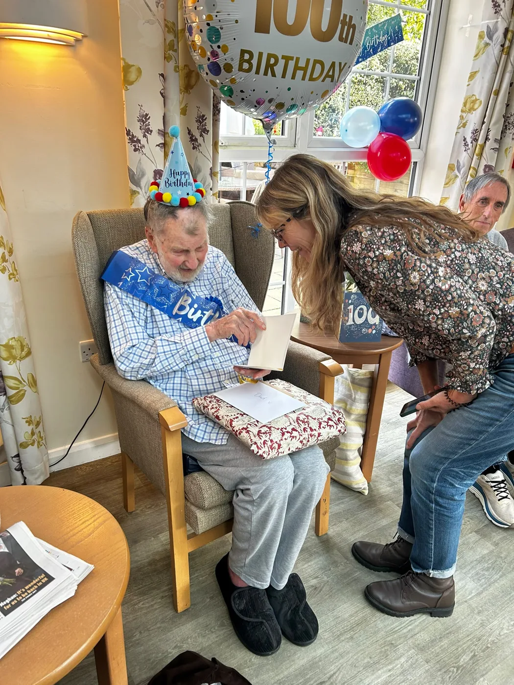 Bill Hopkins in a party hat and blue birthday sash, sitting in an armchair with a card and a wrapped present, as a woman leans in to talk to him under a 100th birthday balloon