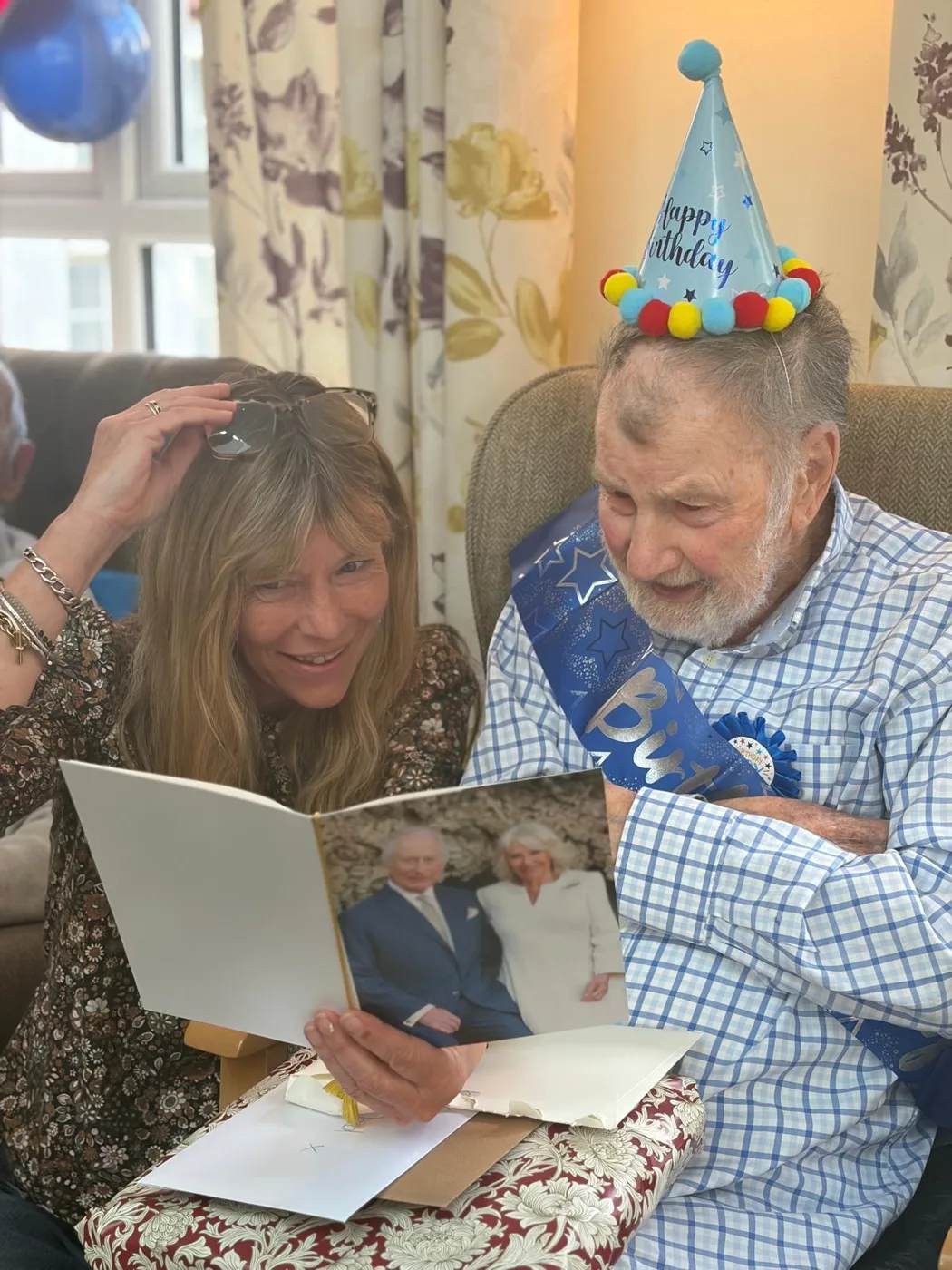 Bill Hopkins in a party hat and birthday sash, smiling as he and a woman read a card with a photograph of the King and Queen on the front