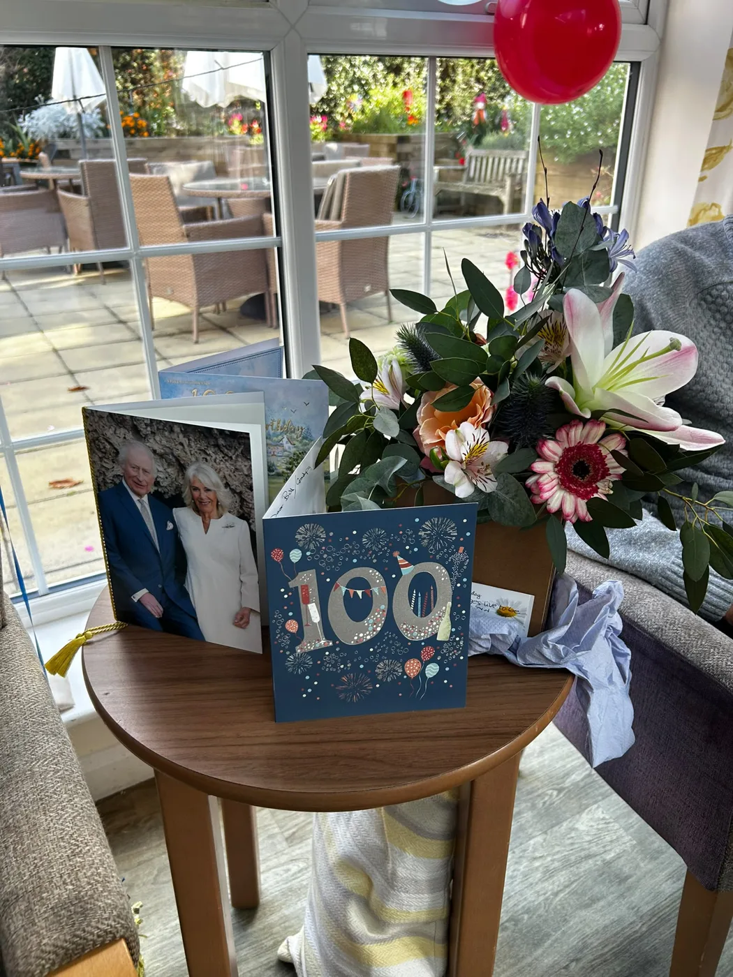Birthday cards, including one with a large 100 on it and one with a photograph of the King and Queen, beside a vase of lilies and gerberas on a small table by the conservatory windows