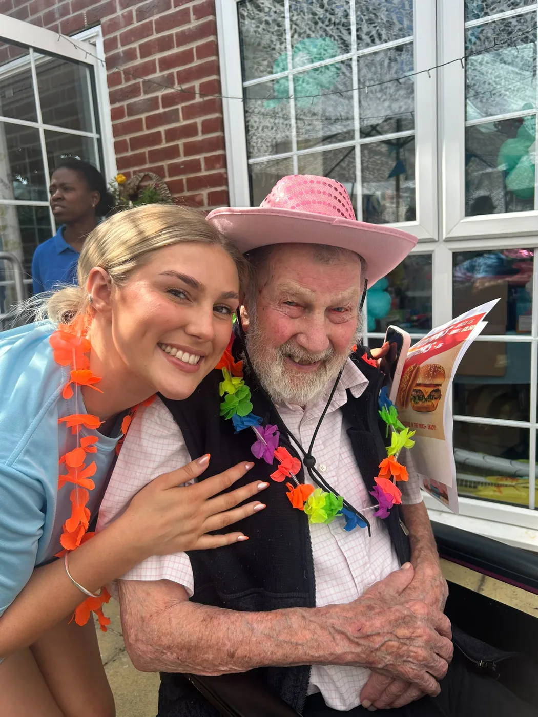 Bill Hopkins in a pink cowboy hat and a rainbow flower garland, smiling beside a young woman outside the home