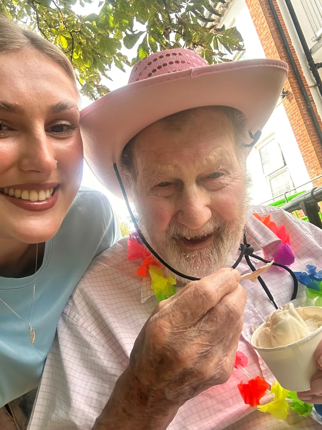 Bill Hopkins in a pink cowboy hat and flower garland eating an ice cream, with a young woman beside him
