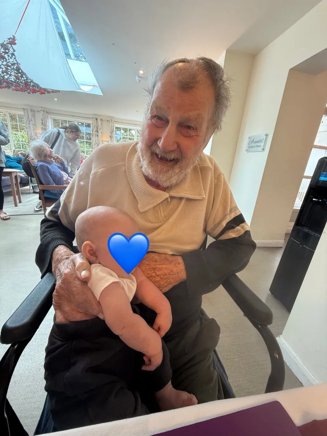 Bill Hopkins smiling as he holds a baby on his lap in the lounge at Downs House; the baby's face is covered with a blue heart