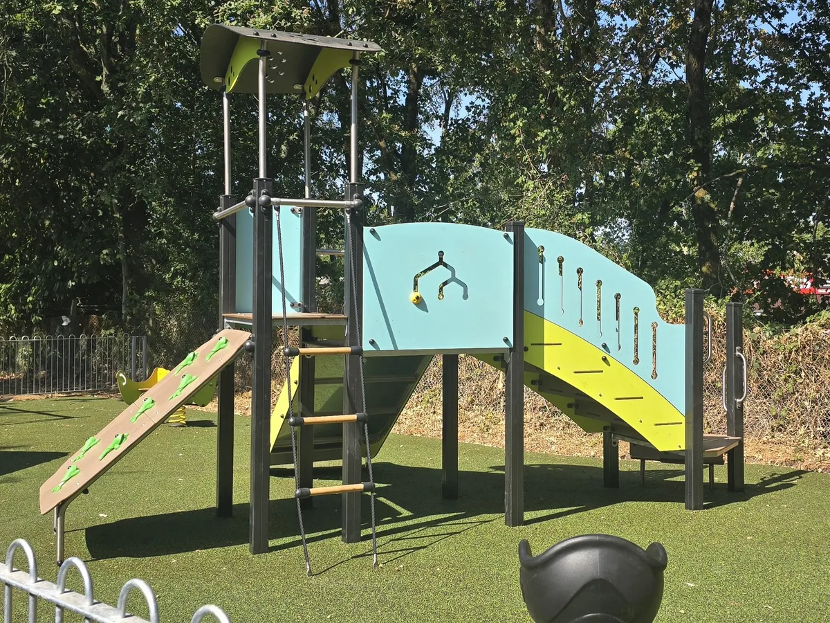 A new climbing frame with a pale blue and yellow slide, a climbing ramp and rope ladder on green safety surfacing, with trees behind.