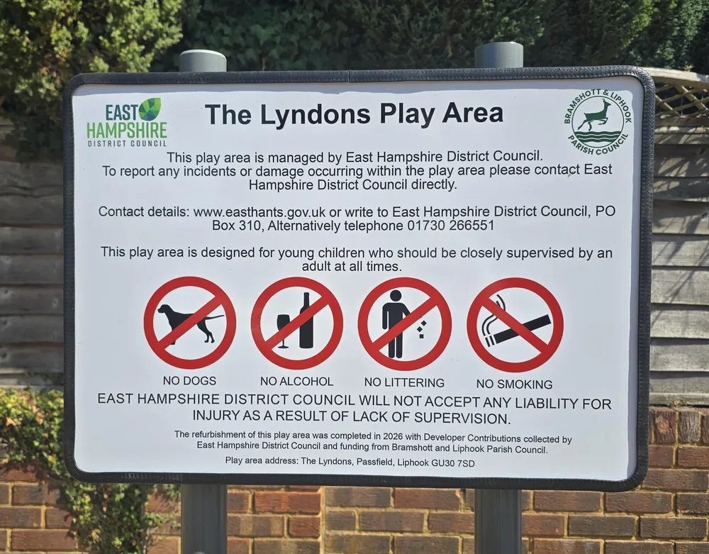 The Lyndons Play Area sign: managed by East Hampshire District Council, report incidents or damage to the council on 01730 266551, designed for young children who should be closely supervised by an adult, no dogs, no alcohol, no littering, no smoking. Refurbished in 2026 with developer contributions and funding from Bramshott and Liphook Parish Council. Address: The Lyndons, Passfield, Liphook GU30 7SD.