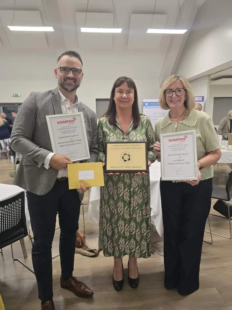 Three people from Bramshott and Liphook standing in a function room, holding the Village of the Year plaque, a joint winner certificate and a runner-up certificate for Best Community Event.