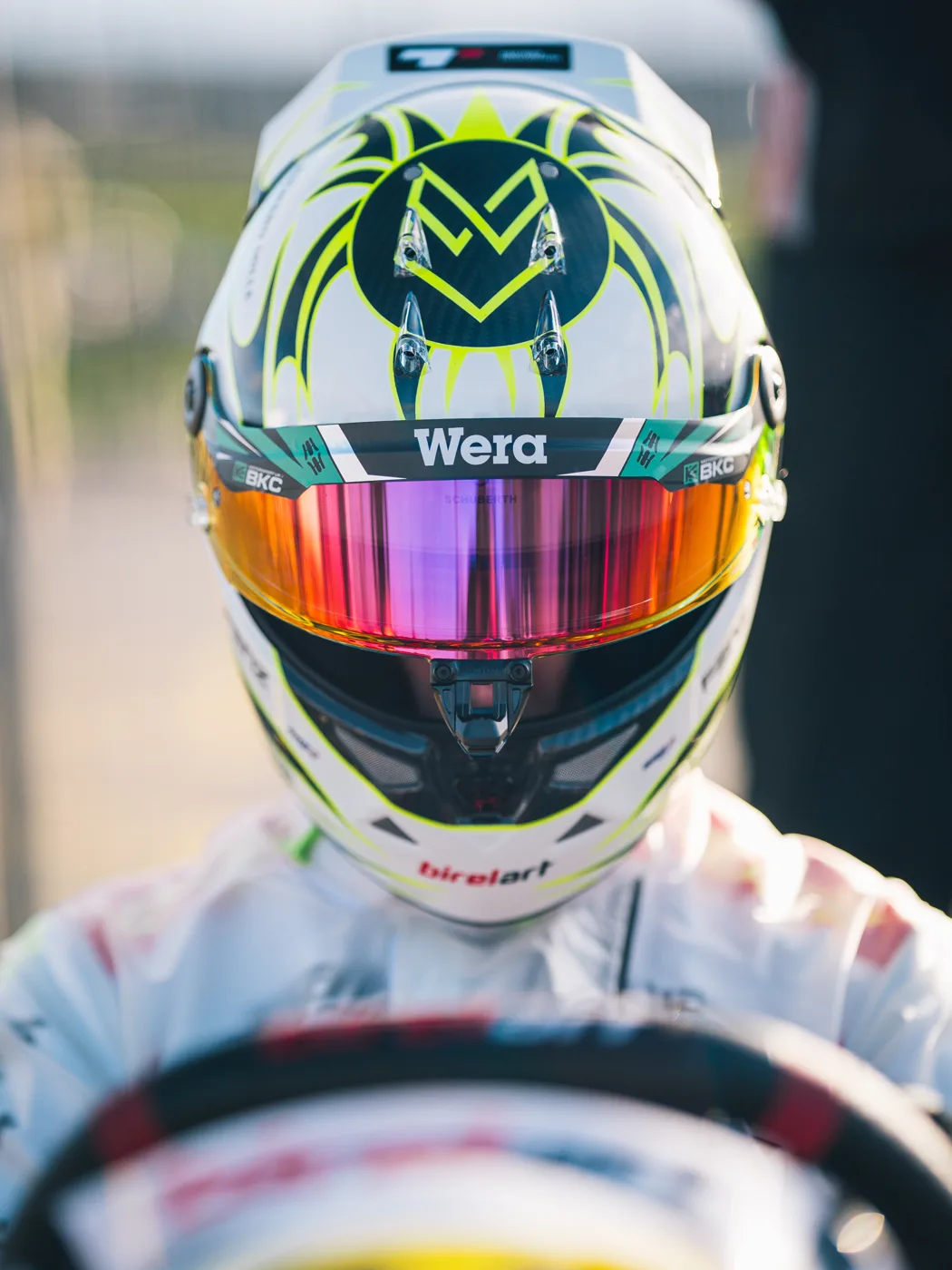 A racing driver seen from the front in a white helmet with yellow and black markings, a Wera visor strip and a mirrored pink and orange visor, hands on a kart steering wheel.