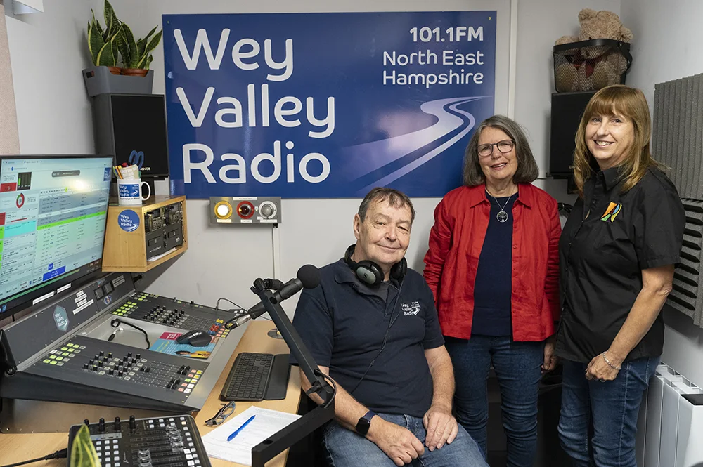 A presenter seated at a radio mixing desk with two women standing beside him, under a large blue Wey Valley Radio 101.1FM North East Hampshire sign in a small studio.